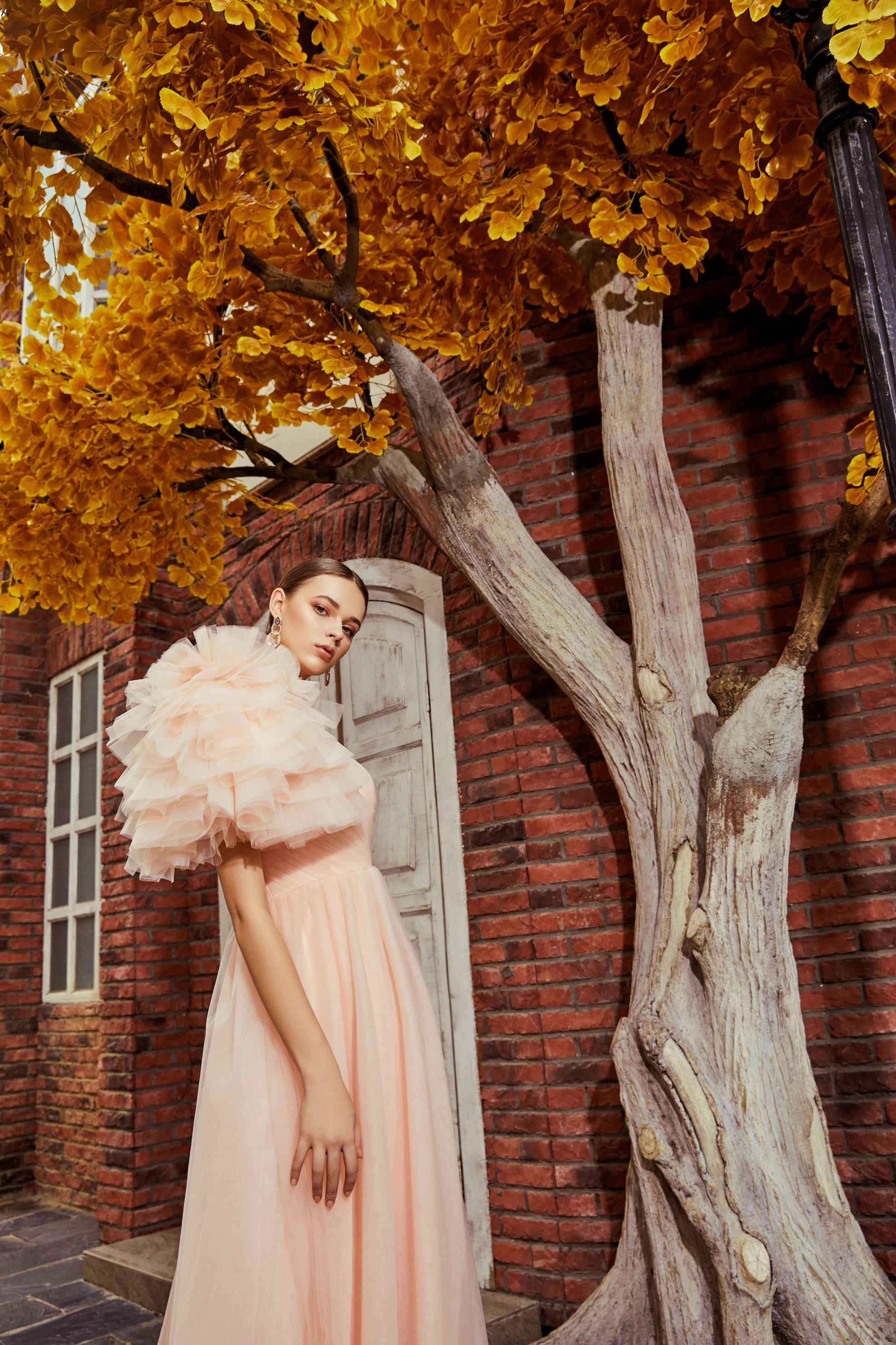 Woman in a light pink dress standing under a tree with autumn leaves in front of a brick building.